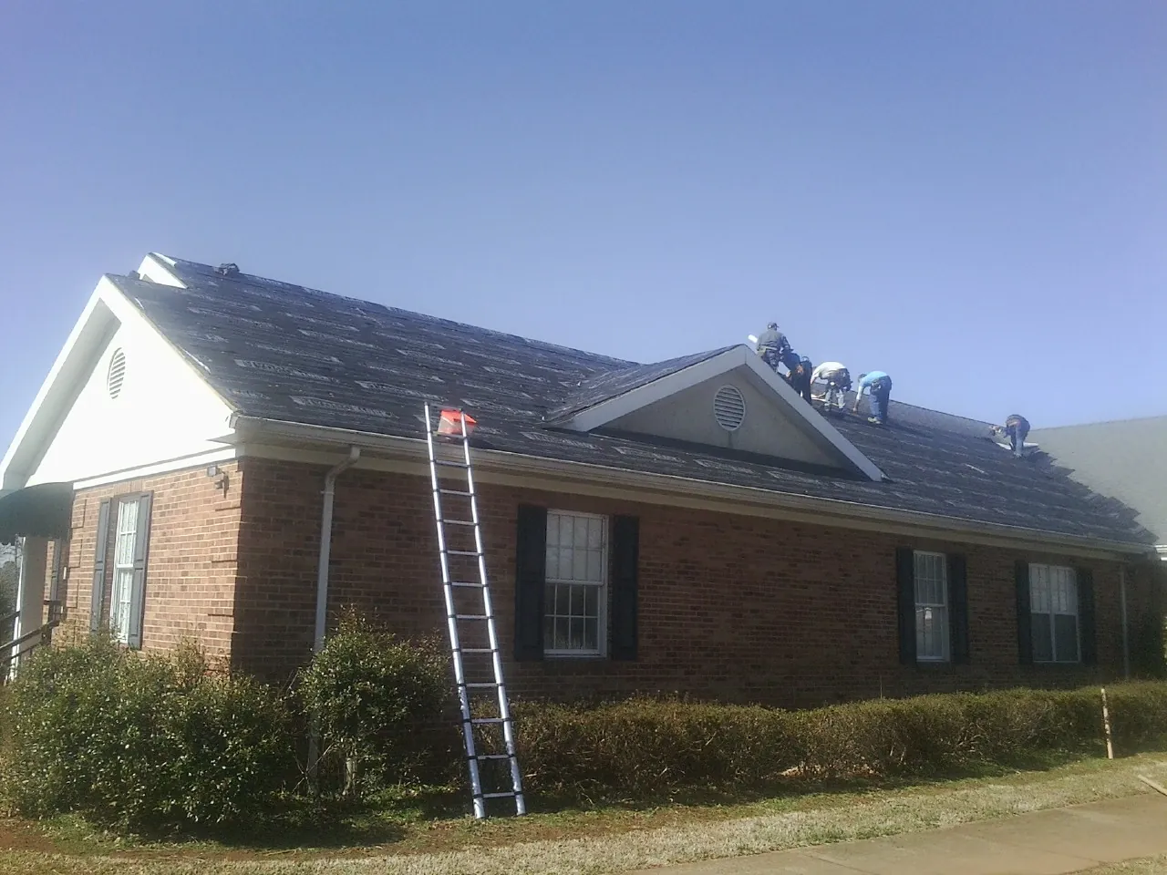 Crew tearing shingles off a brick ranch roof with a ladder set — Metal Roof Repair work by our crew in Heron
