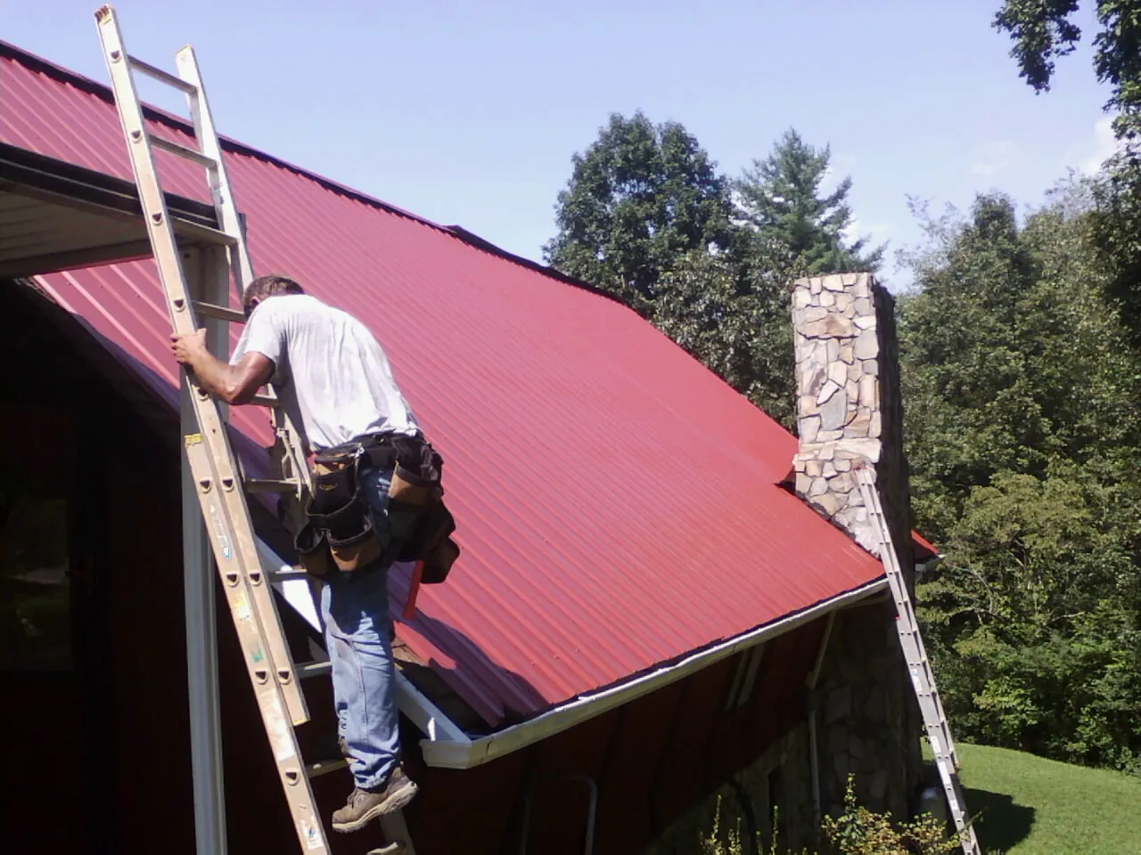 Roofer on a ladder beside a red metal roof and stone chimney — Soffit & Fascia Repair by Roofing Brothers in Heron